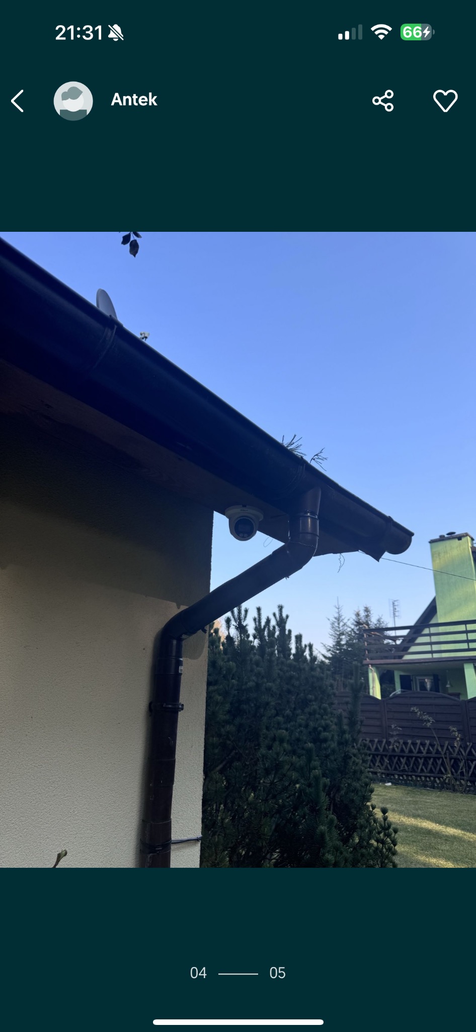 Kamera monitoringu zamontowana pod dachem budynku z brązową rynną. Widoczne krzewy i fragment domu w tle, błękitne niebo.