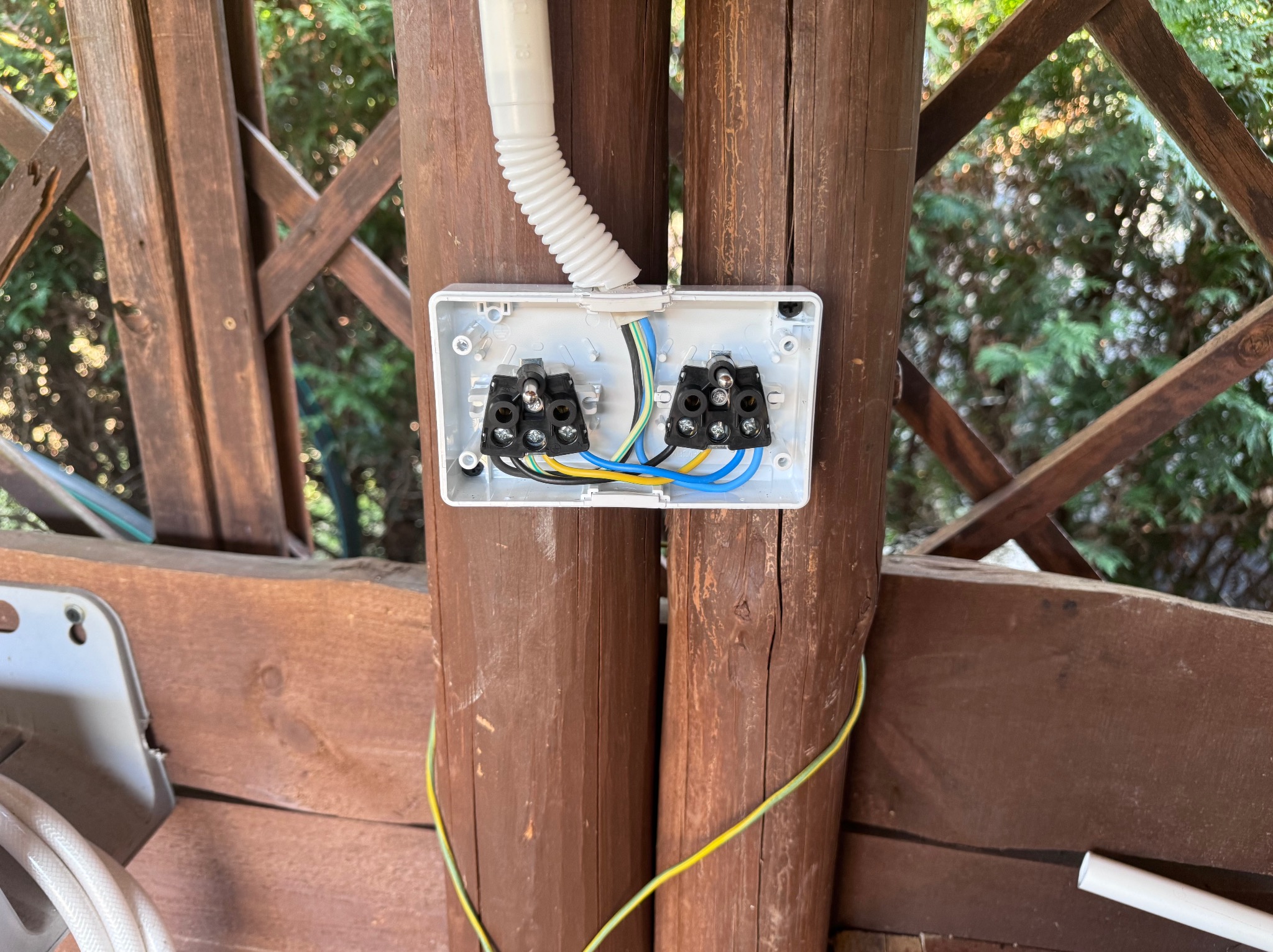 Otwarta puszka elektryczna z podłączonymi przewodami na drewnianej konstrukcji altany. Widoczne złącza i fragment peszla ochronnego.