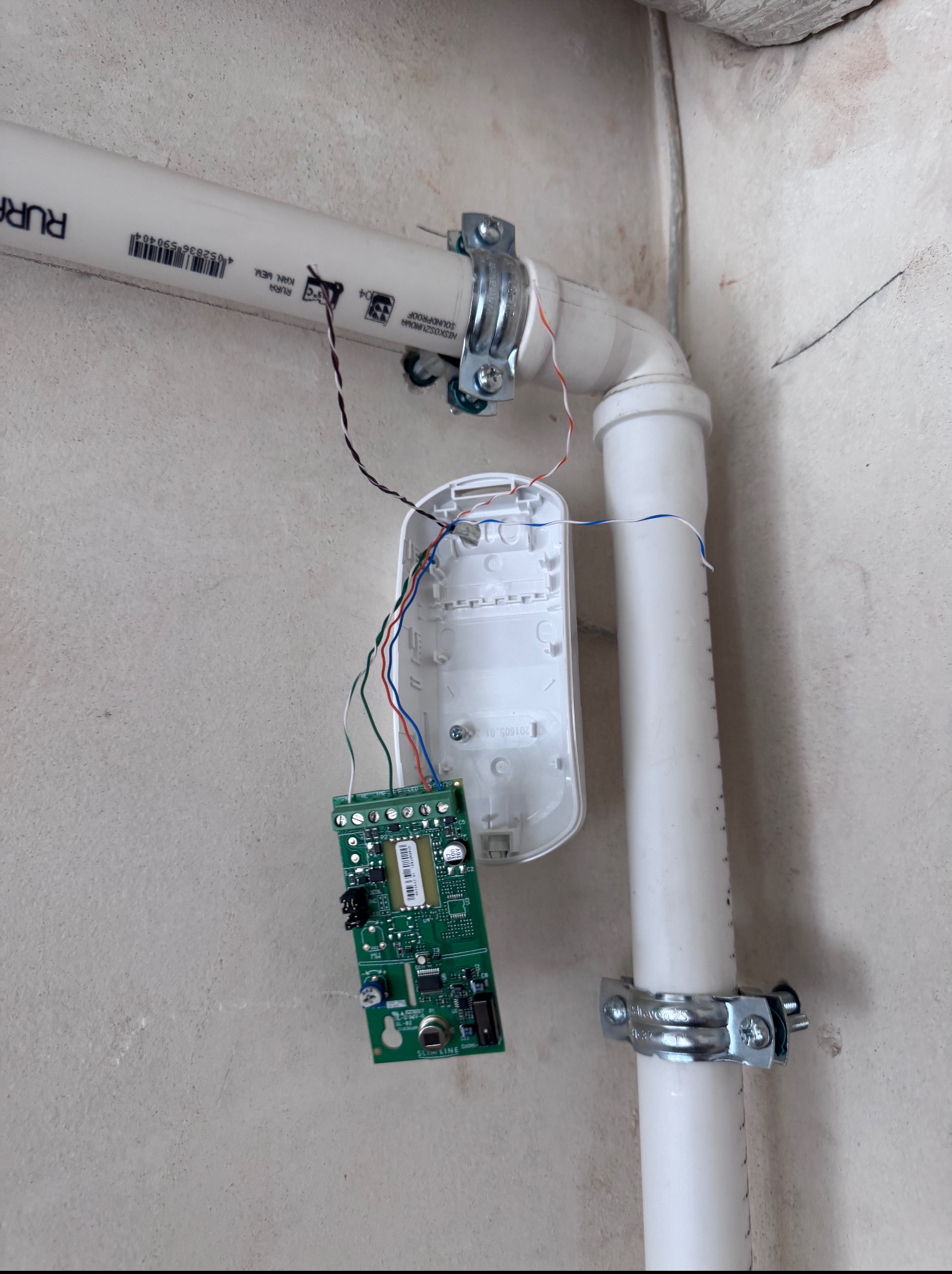 Rozmontowany czujnik alarmowy z widoczną płytką drukowaną, podłączony do przewodów, mocowany do ściany za pomocą rury i metalowych uchwytów.