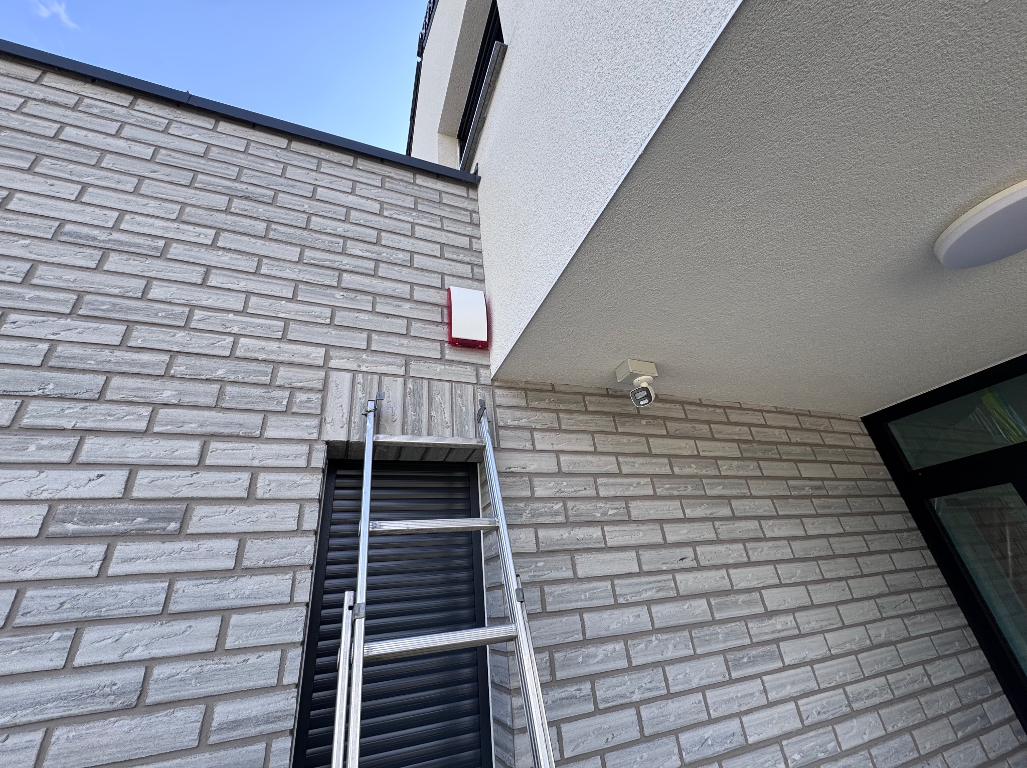 Biała kamera monitoringu i sygnalizator alarmowy na elewacji z cegły. Aluminiowa drabina oparta o ścianę. Widok z dołu na budynek.