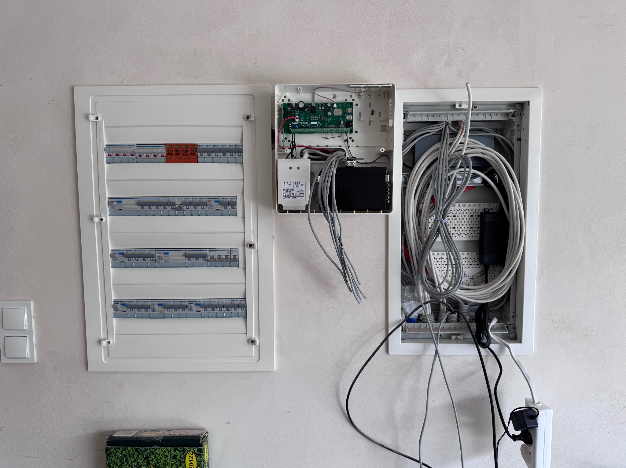 Rozdzielnica elektryczna z bezpiecznikami i okablowaniem w białej obudowie na ścianie. Widoczna instalacja alarmowa i zasilacz. Częściowo otwarte skrzynki.