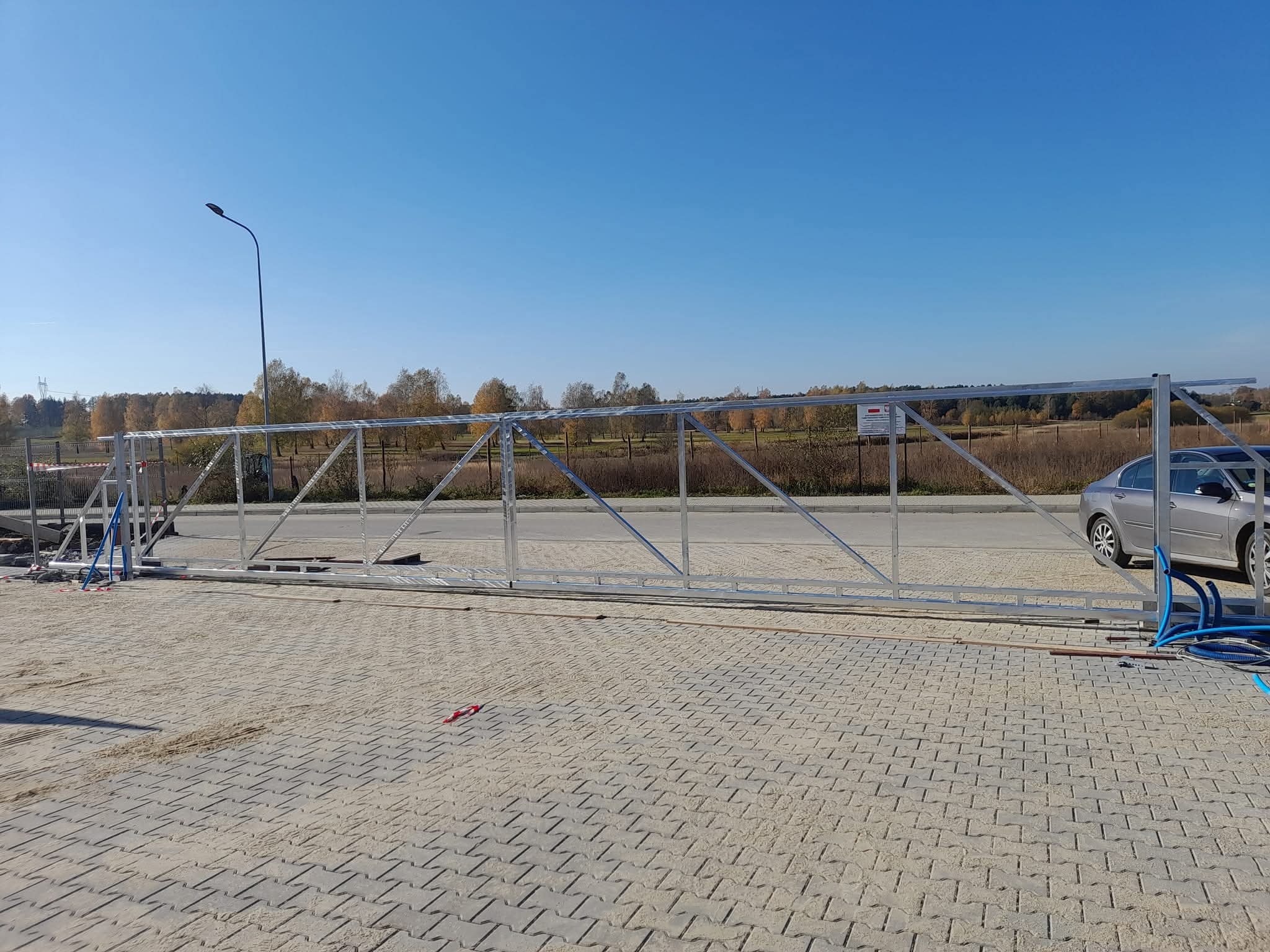 Srebrna, metalowa konstrukcja bramy przesuwnej, jeszcze bez wypełnienia, leżąca na brukowanym podłożu. W tle drzewa i błękitne niebo.