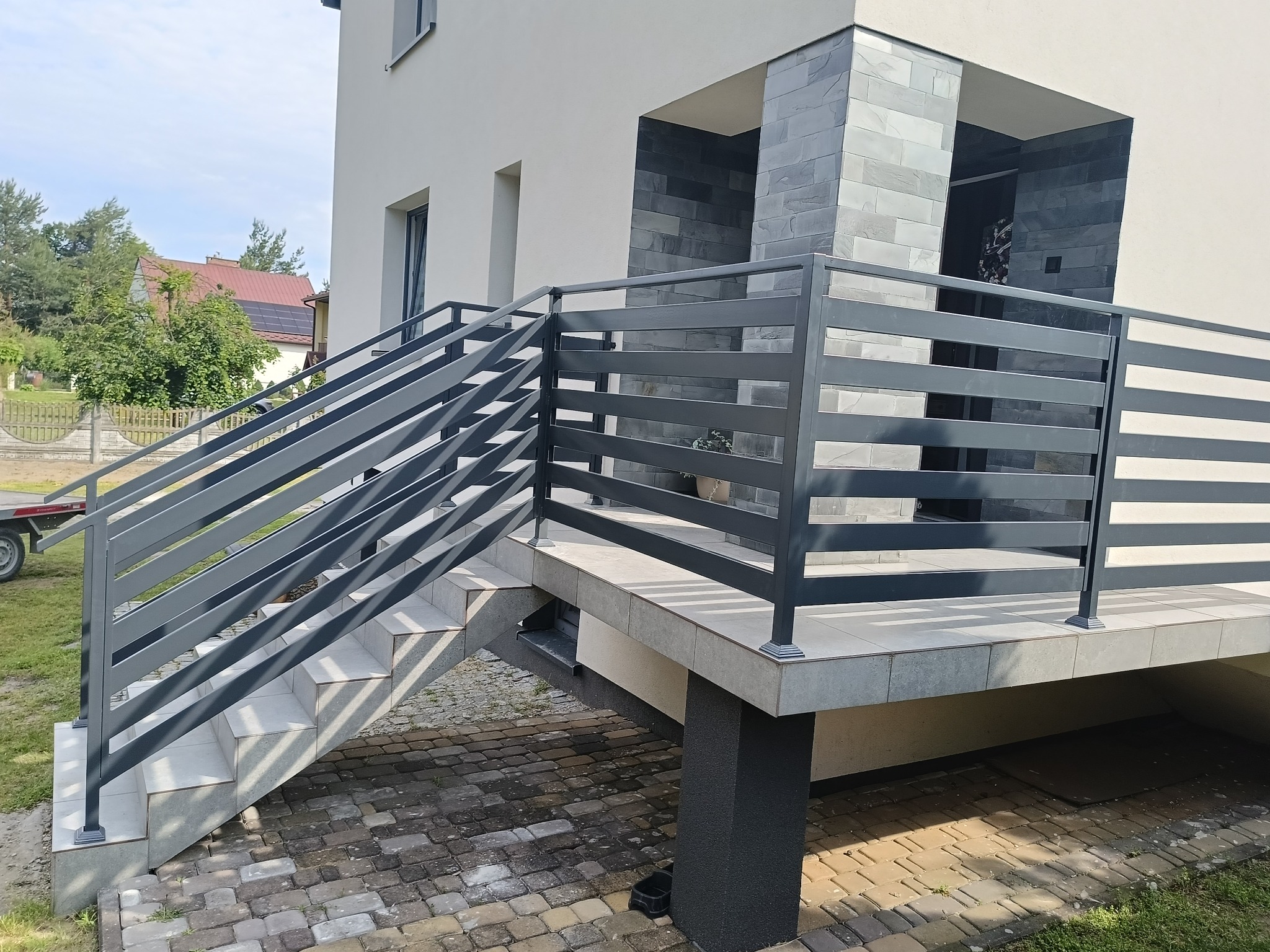 Nowoczesne schody zewnętrzne z szarą, poziomą balustradą, prowadzące do wejścia domu z elementami z szarego kamienia. Widok na kostkę brukową i fragment trawnika.