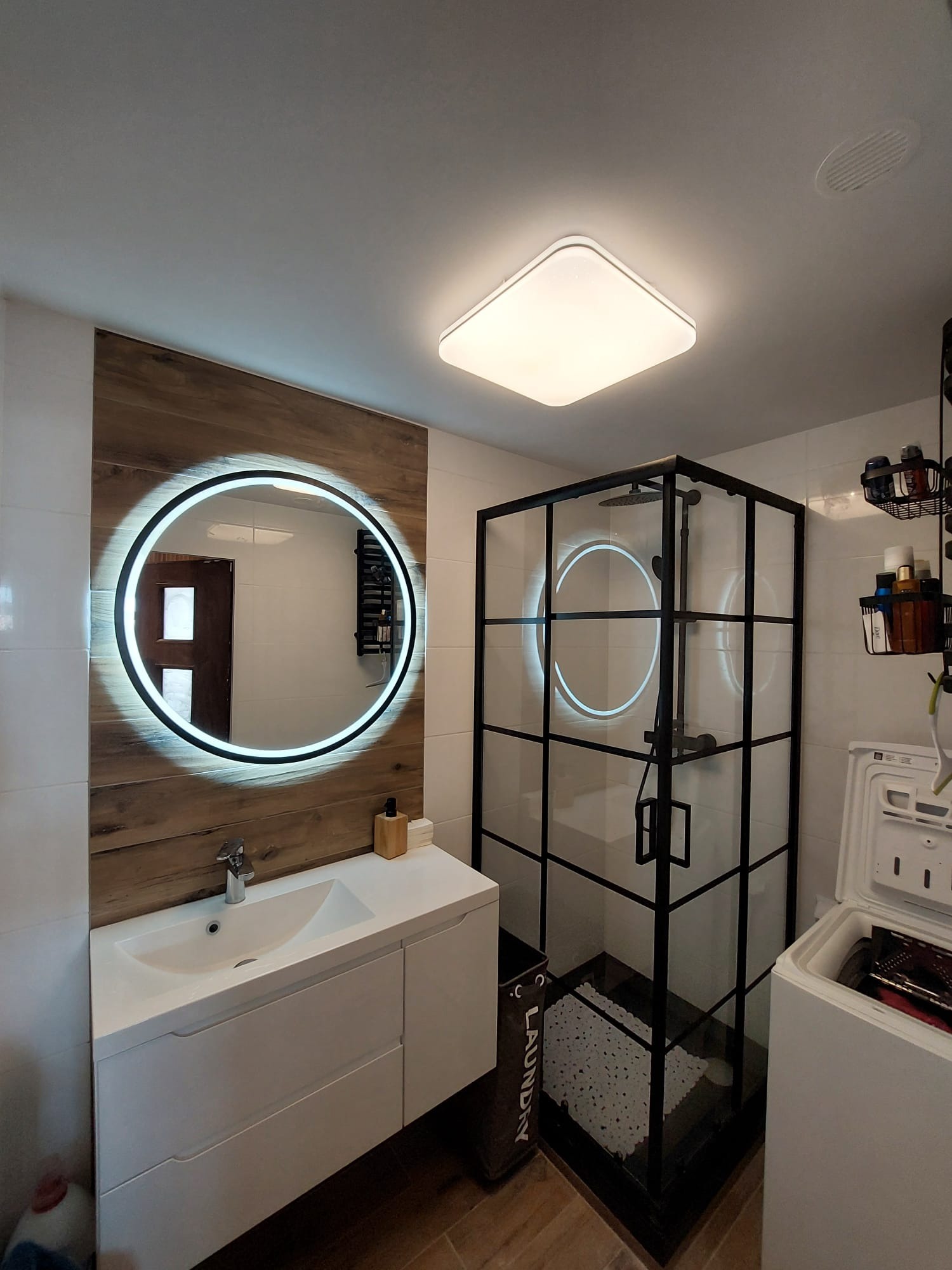 Nowoczesna łazienka z kabiną prysznicową w czarnej ramie, okrągłym lustrem LED i białą szafką z umywalką. Widoczna pralka i kosz na pranie.