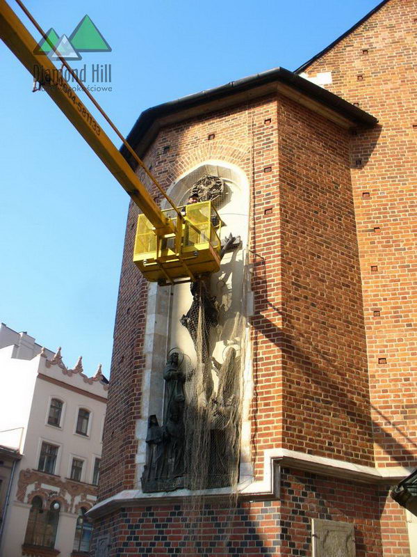 Prace wysokościowe przy renowacji elewacji zabytkowej kamienicy z cegły, widoczna platforma robocza i rzeźba religijna na ścianie budynku.