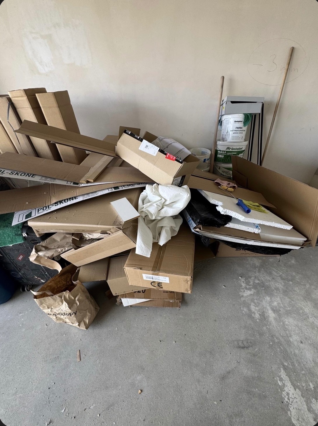 Stos kartonów, gruzu i farb na podłodze w pomieszczeniu. Widoczne puszki farb Greinplast i fragmenty materiałów budowlanych. Ściana z notatkami.