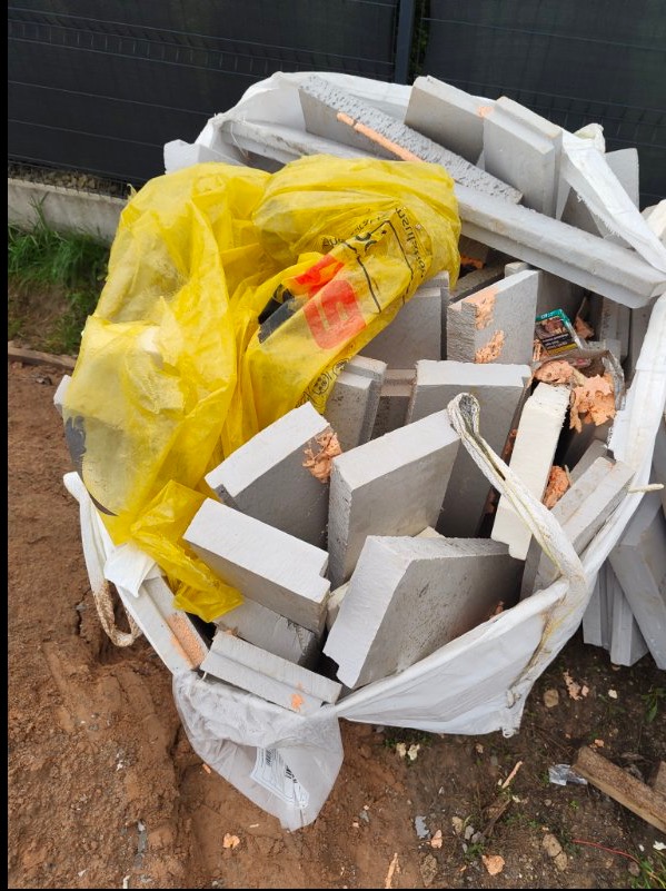 Worek z gruzem pobudowlanym, żółta torba foliowa, fragmenty styropianu i cegieł. Widoczne ślady zaprawy. Teren budowy w tle.
