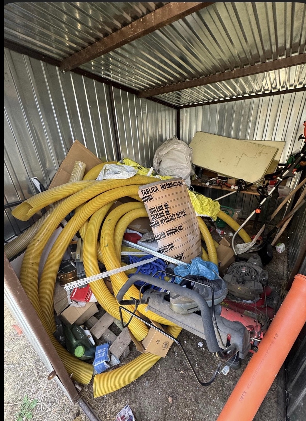 Zabałaganiony garaż blaszany z żółtymi rurami, narzędziami ogrodniczymi i tablicą informacyjną budowy. Widok z góry na nieporządek wewnątrz.