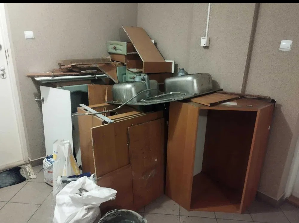 Stos zdemontowanych mebli kuchennych, w tym szafki, blaty i dwie metalowe umywalki, zgromadzony w rogu pomieszczenia. Widoczne worki i wiadro na podłodze.
