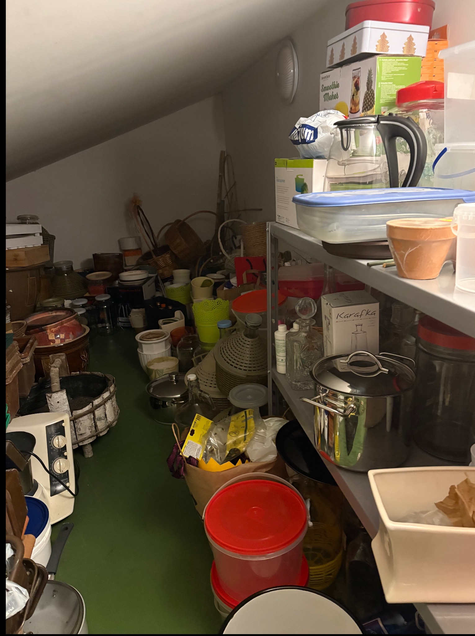 Zagracone poddasze ze stertą różnorodnych przedmiotów, od doniczek i koszy po sprzęt AGD i pudełka. Dominują przedmioty codziennego użytku, tworząc wrażenie chaosu.