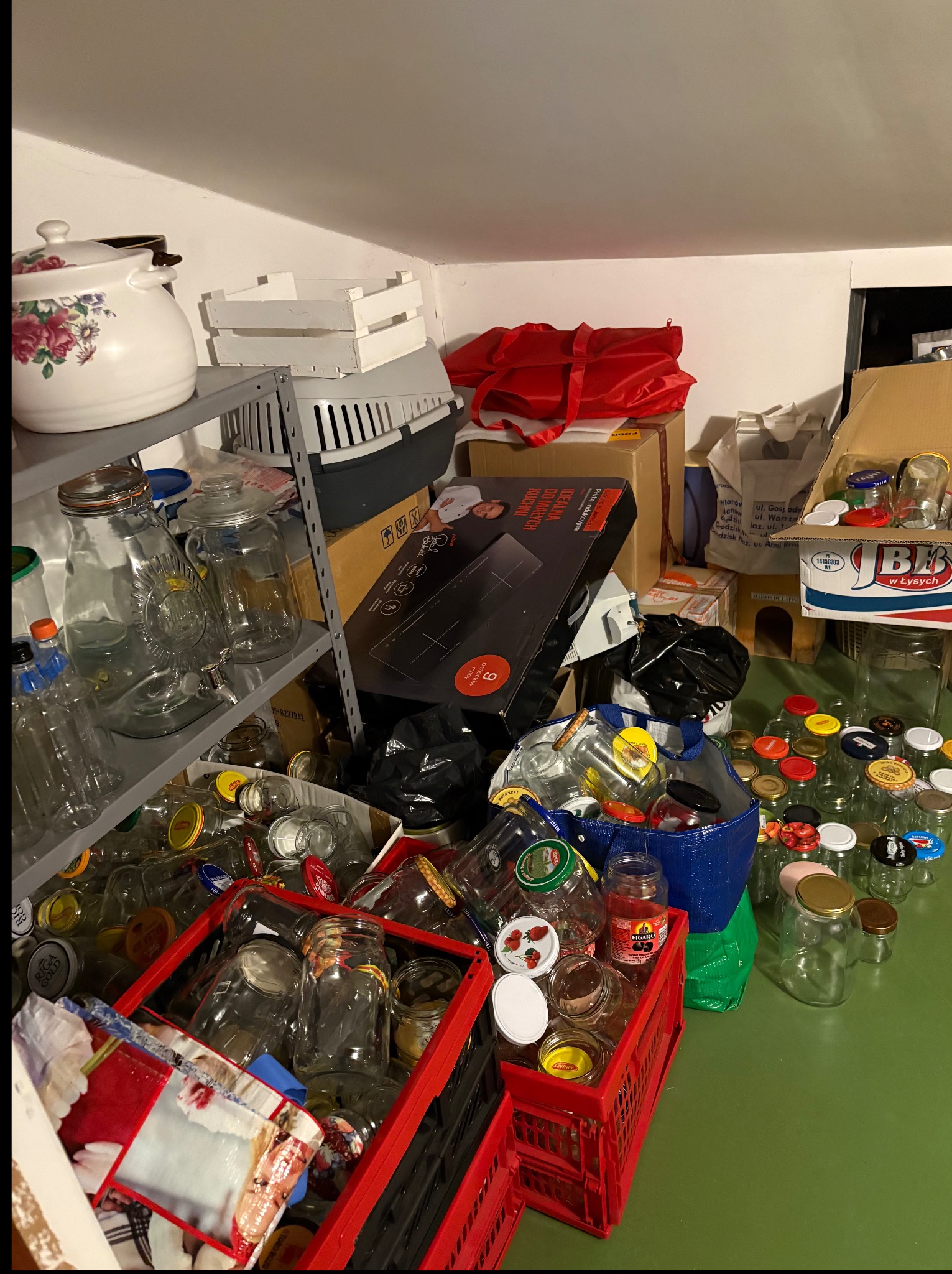 Zagracone pomieszczenie: słoiki w skrzynkach, torbach, pudełkach, na półkach. Widoczny regał, pudełka, torby, skrzynki. Chaos i nieporządek w pomieszczeniu.