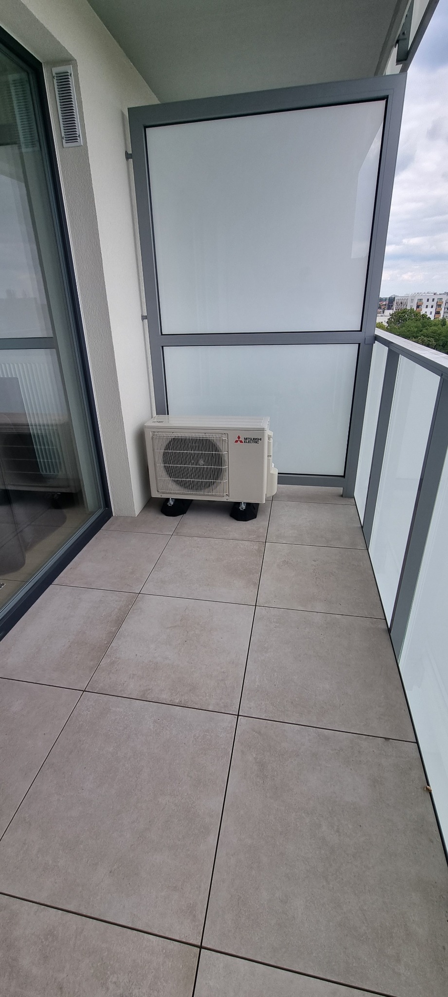 Kompaktowa klimatyzacja Mitsubishi Electric na balkonie nowoczesnego apartamentu, ustawiona na podkładkach antywibracyjnych. Widok na miasto w tle.