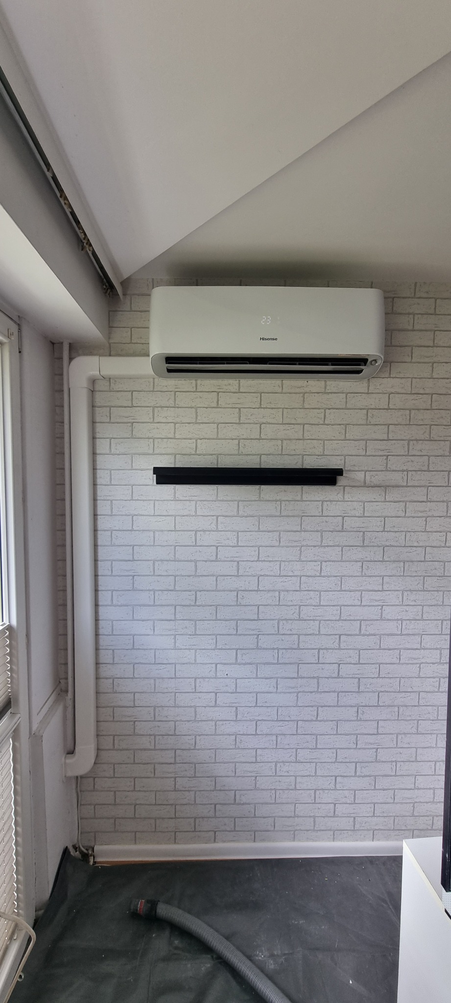 Biała klimatyzacja Hisense zamontowana na ścianie z białej cegły. Widoczna rura instalacyjna, czarna półka i wąż odkurzacza na podłodze.