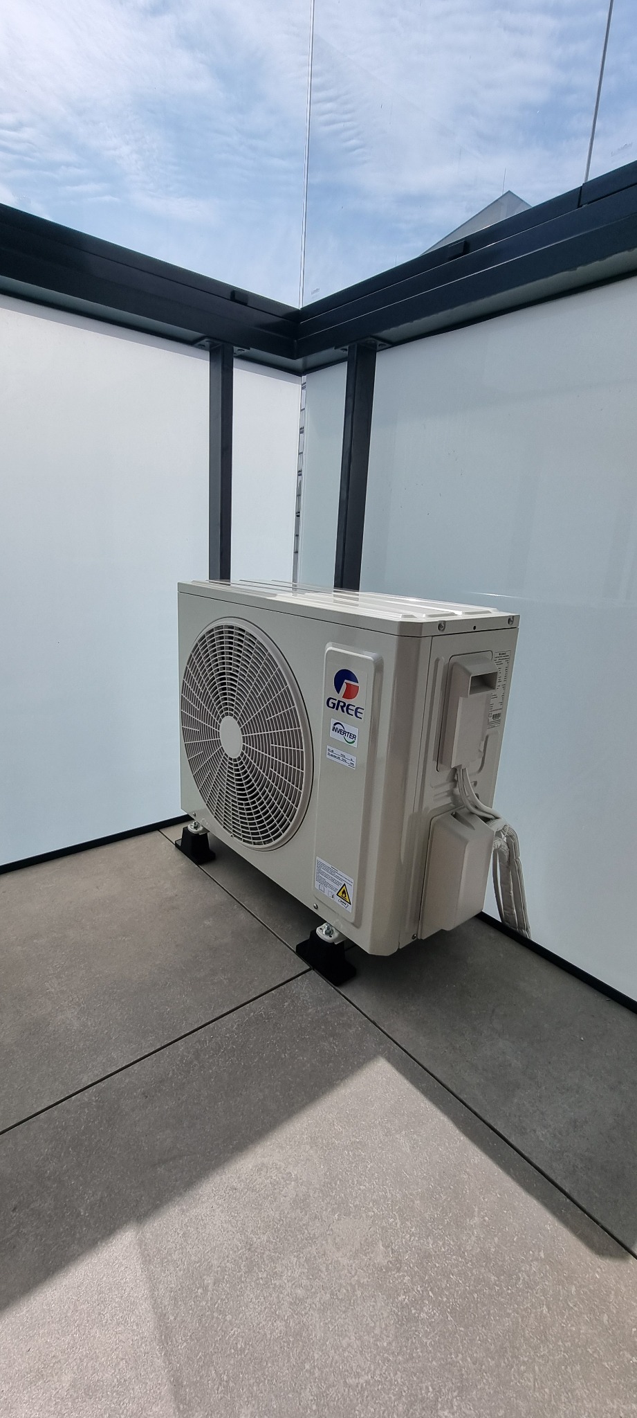 Nowoczesny klimatyzator Gree Inverter zamontowany na szarym tarasie, widok z boku na tle białej ściany i szklanego zadaszenia.