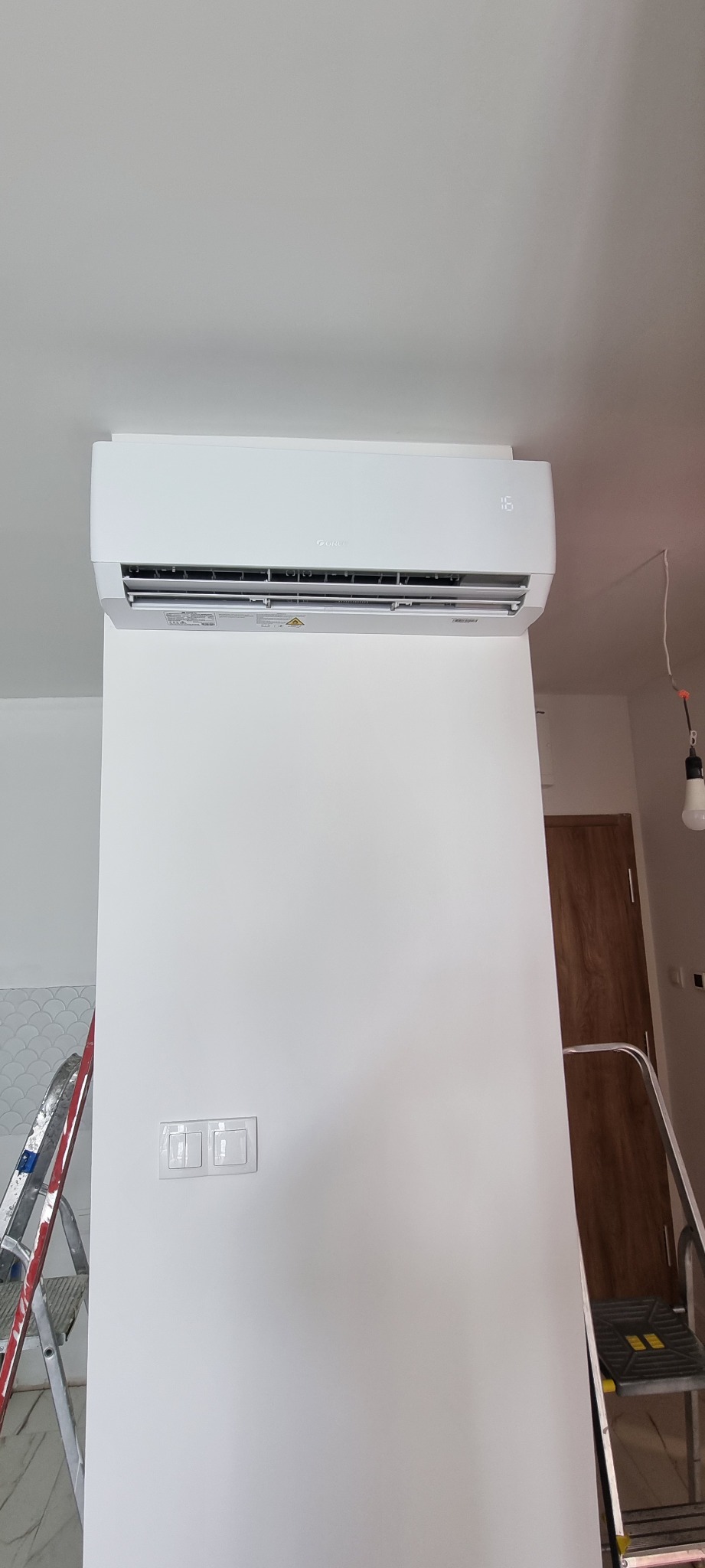 Biały klimatyzator ścienny marki Gree zamontowany na ścianie w jasnym wnętrzu, widoczne dwa metalowe stojaki i włączniki światła.