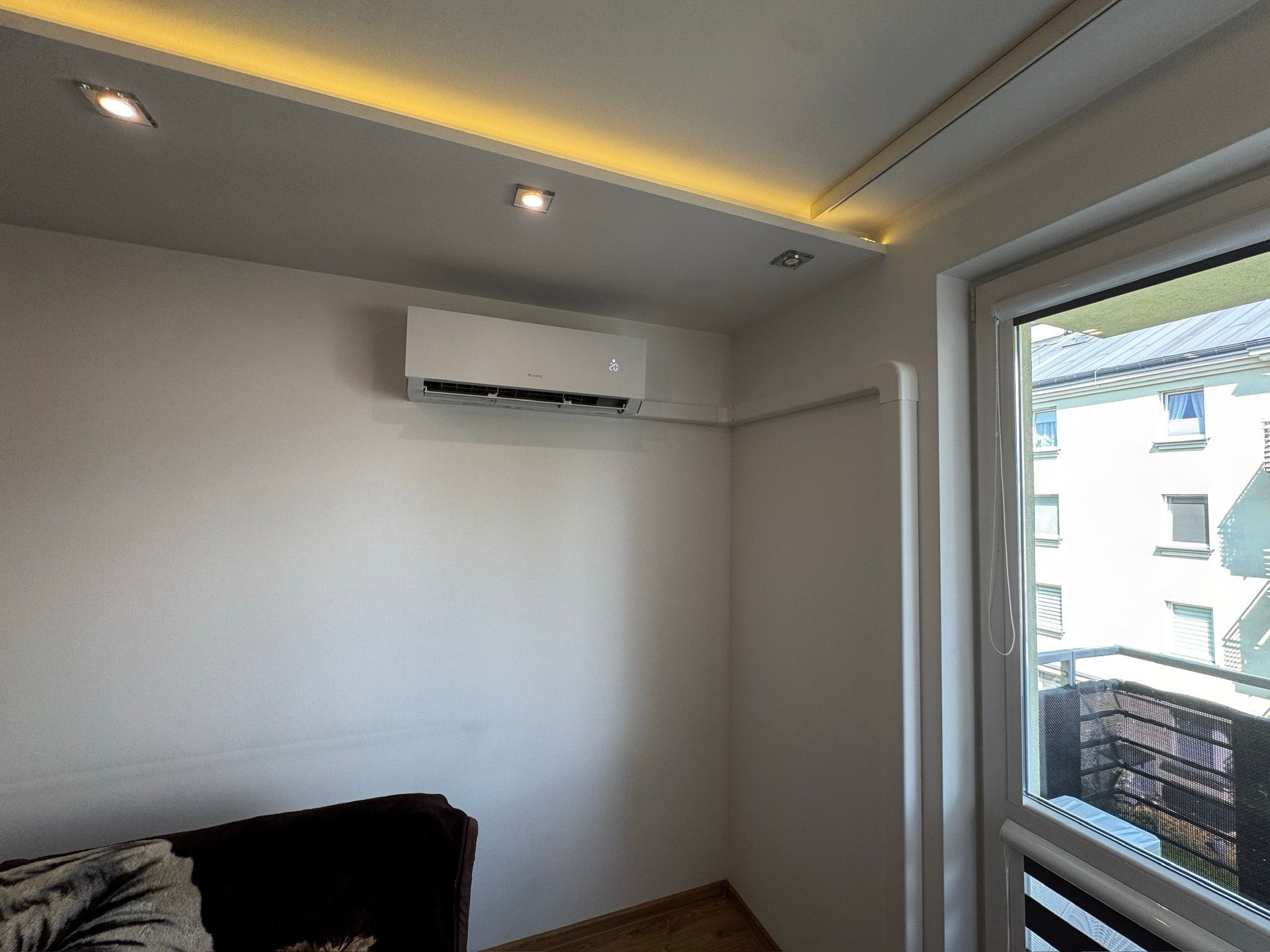 Biały klimatyzator z widocznym wyświetlaczem temperatury zamontowany w pokoju z oświetleniem sufitowym LED i oknem balkonowym.
