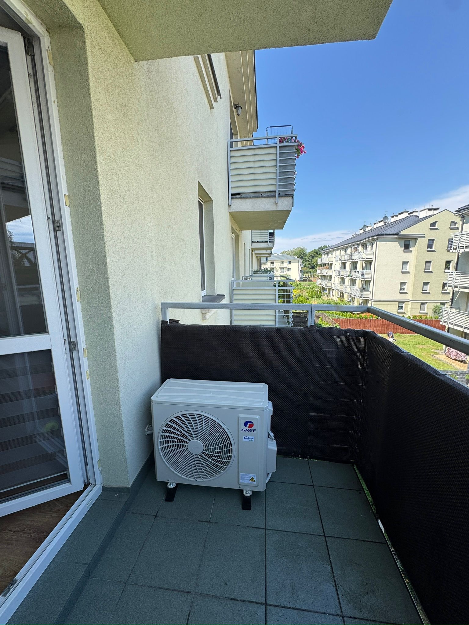 Biała jednostka zewnętrzna klimatyzacji Gree na balkonie bloku mieszkalnego z widokiem na inne balkony i budynki w słoneczny dzień.