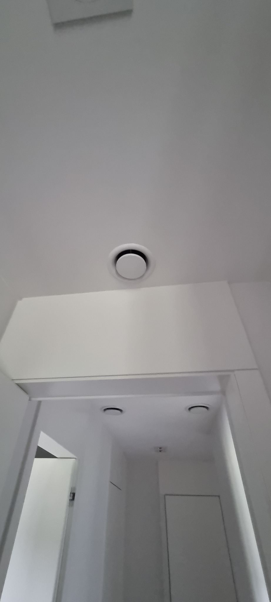 Biały sufit z okrągłym nawiewnikiem klimatyzacji i kratką wentylacyjną. Widok z dołu w kierunku korytarza.