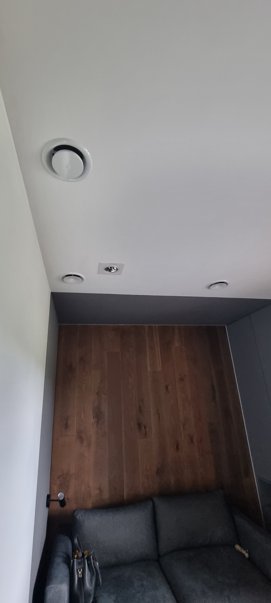 Nowoczesne nawiewniki klimatyzacyjne sufitowe, okrągłe i kwadratowe, wkomponowane w biały sufit nad drewnianą ścianą i szarą sofą, oświetlenie punktowe.