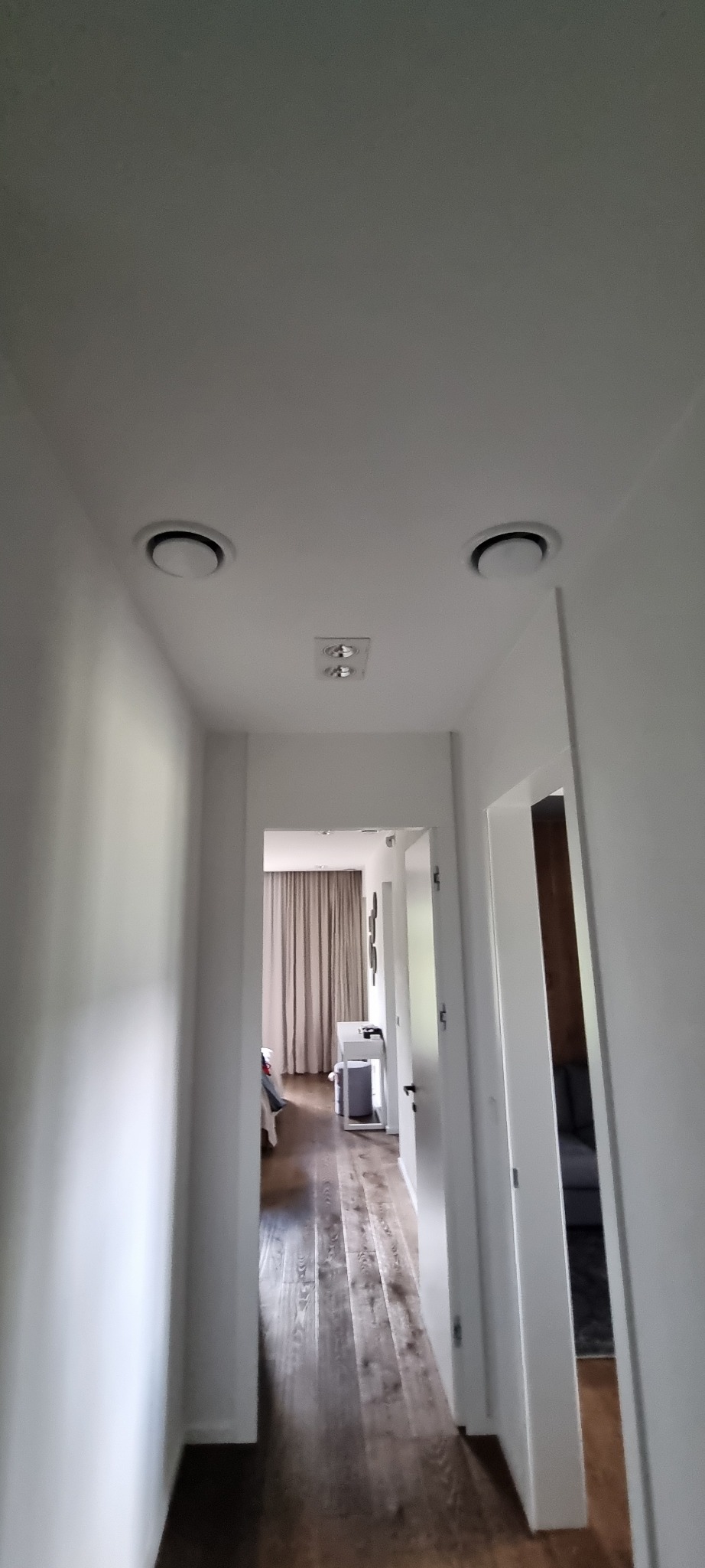 Dwa okrągłe nawiewniki klimatyzacji sufitowej w korytarzu z widokiem na pokój. Minimalistyczny design, białe ściany, drewniana podłoga.