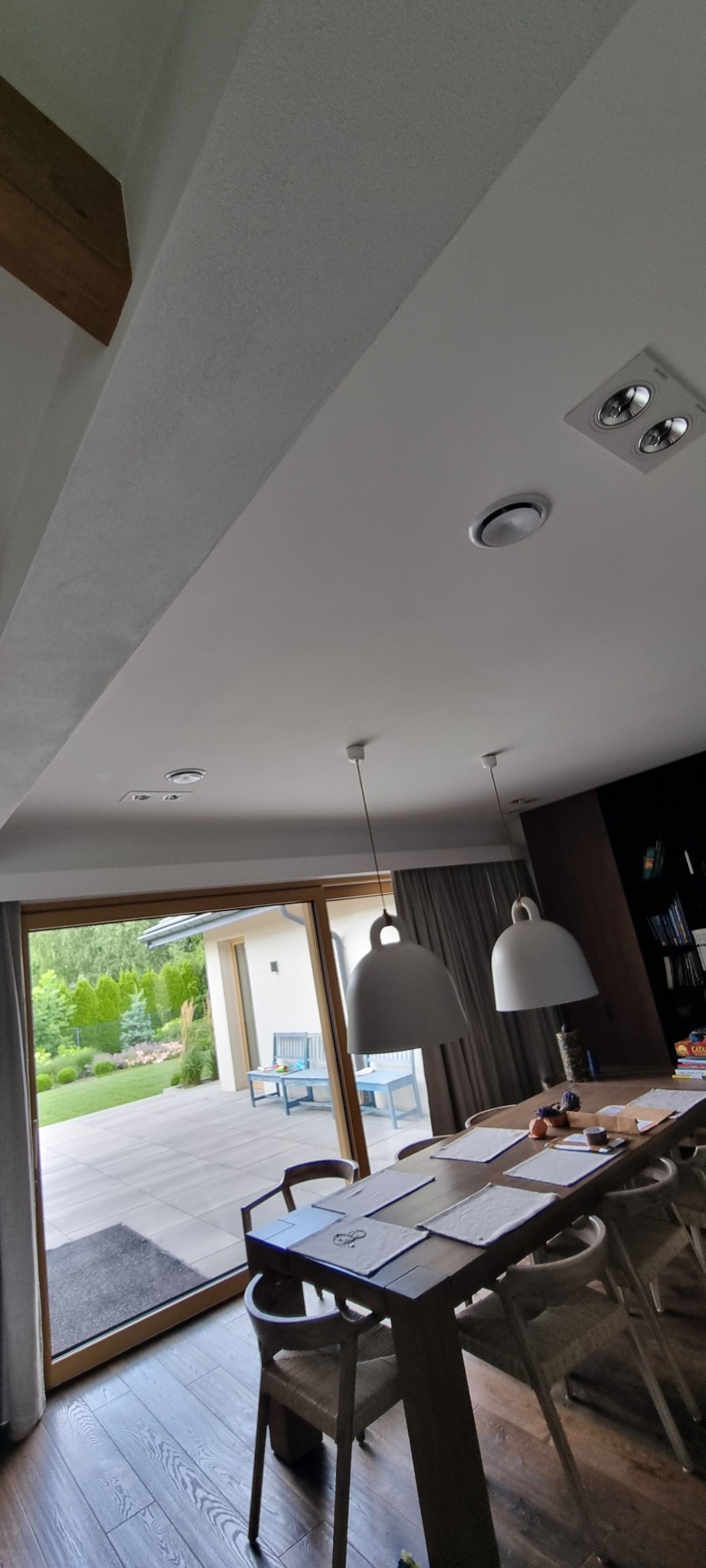 Nowoczesny system wentylacyjny sufitowy z okrągłymi nawiewnikami, widok na taras i ogród przez przesuwne drzwi balkonowe. Oświetlenie: lampy wiszące.