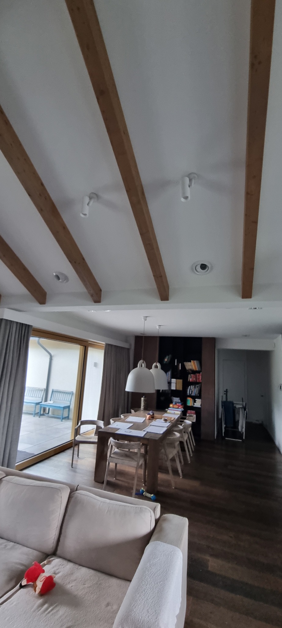 Jasne wnętrze salonu z drewnianymi belkami stropowymi i oświetleniem punktowym. Widoczny stół z krzesłami, sofa i okno z widokiem na zewnątrz.