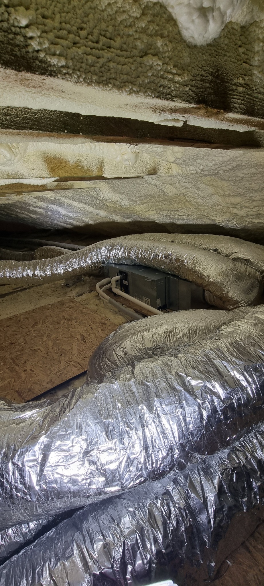 Izolowany strych z widocznymi przewodami wentylacyjnymi i jednostką klimatyzacyjną. Pianka izolacyjna pokrywa krokwie, a przewody wentylacyjne są owinięte folią aluminiową.