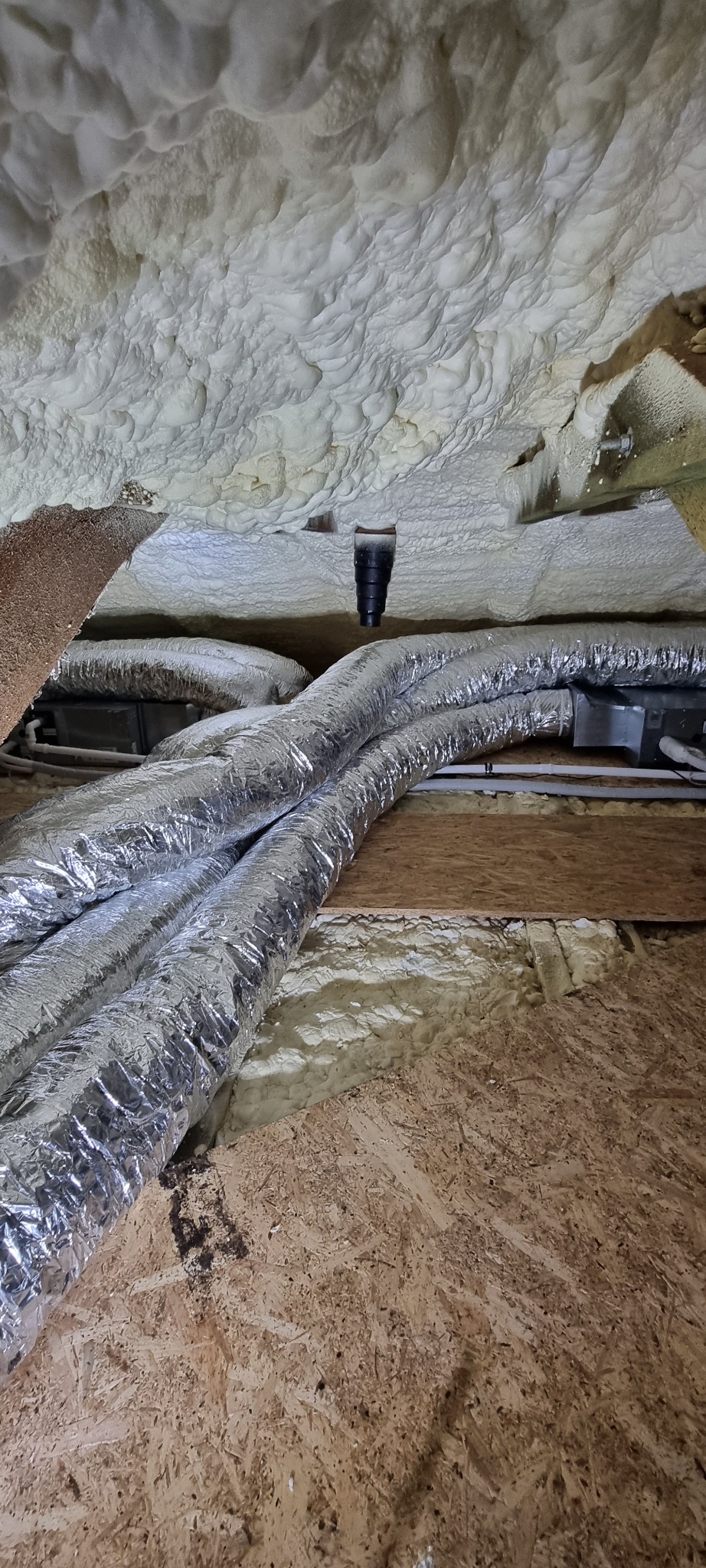 Ocieplenie pianą i srebrne rury wentylacyjne w przestrzeni poddasza w Pruszkowie. Widok z dołu na instalację.