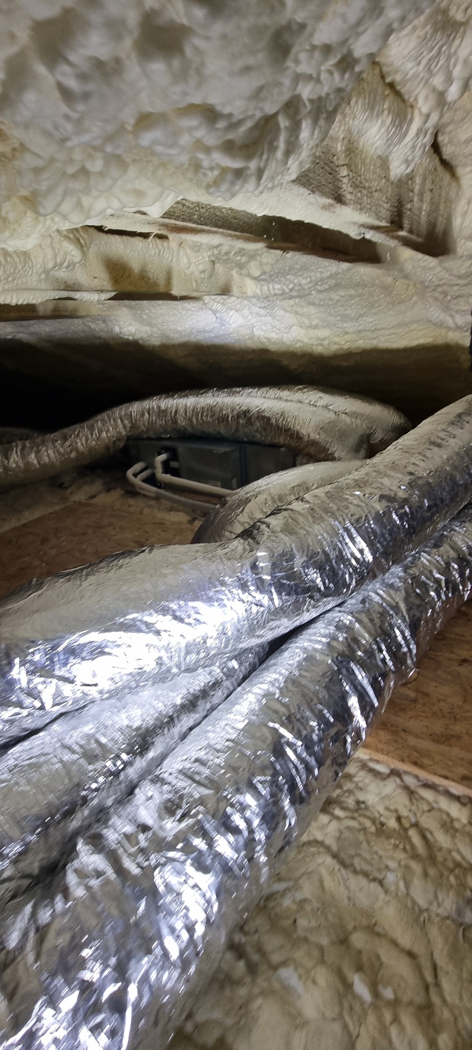 Izolowane przewody wentylacyjne klimatyzacji w przestrzeni poddasza, owinięte folią aluminiową i otoczone pianką izolacyjną. Widoczna skrzynka instalacyjna.