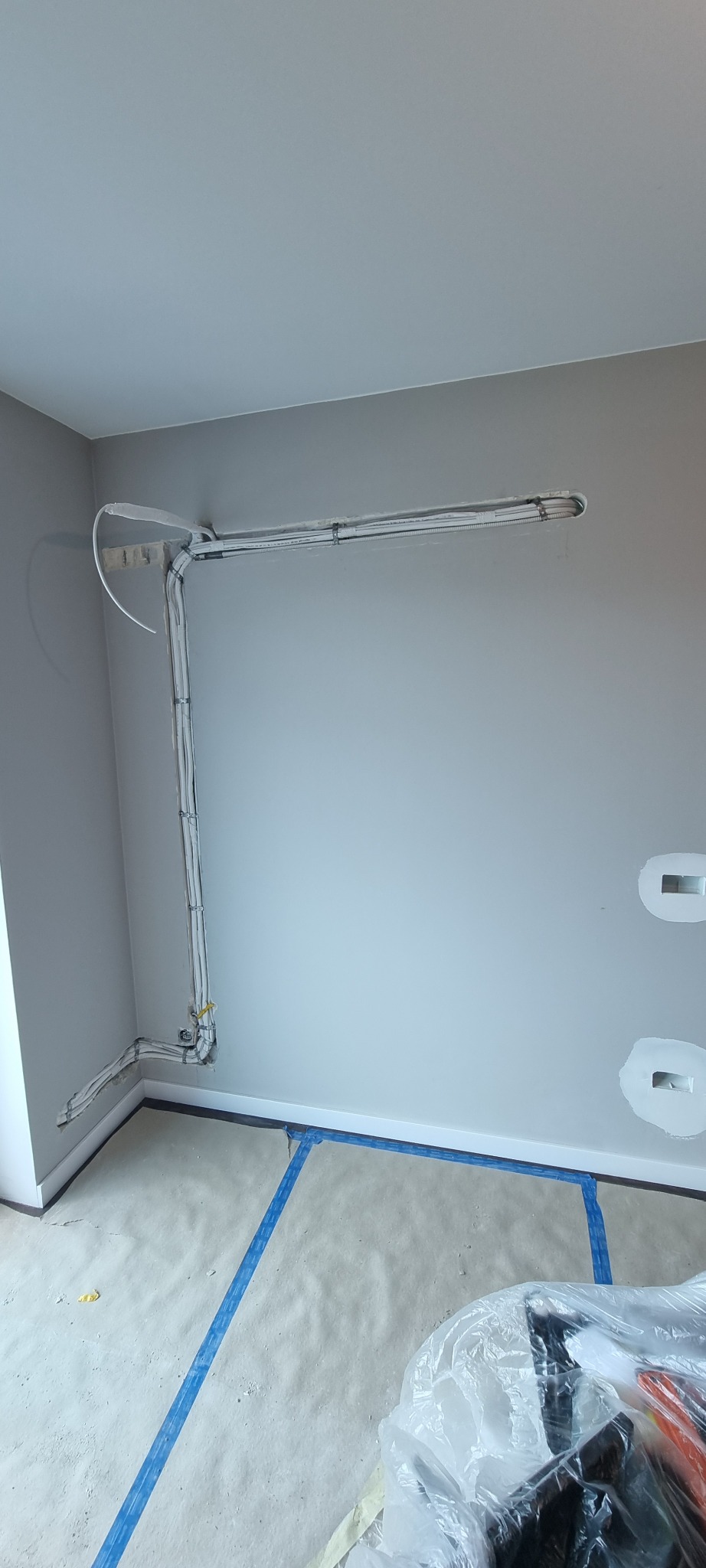 Białe rury instalacji elektrycznej, przymocowane do szarej ściany w nowym budynku, z widocznymi otworami na gniazdka i zabezpieczonym podłożem.