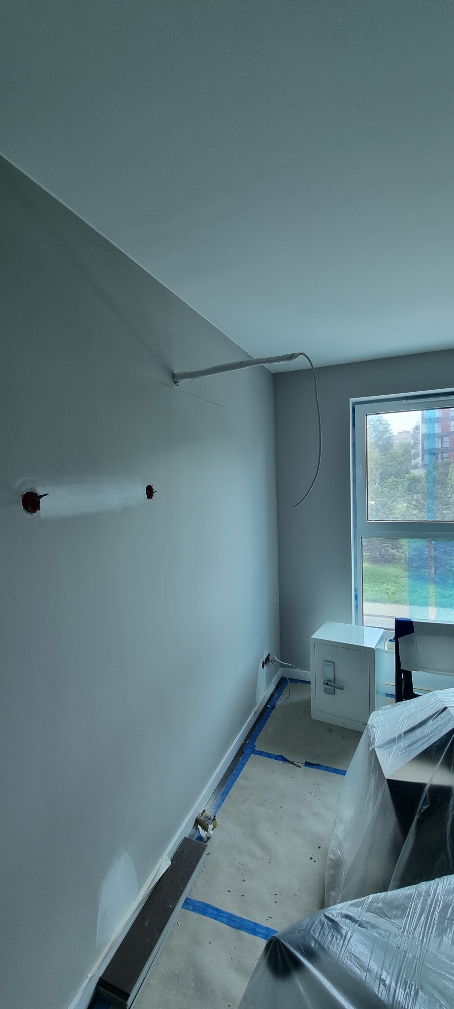 Wnętrze w trakcie instalacji klimatyzacji: widoczne przewody, otwory w ścianie i biała jednostka wewnętrzna przy oknie. Podłoga zabezpieczona niebieską taśmą.