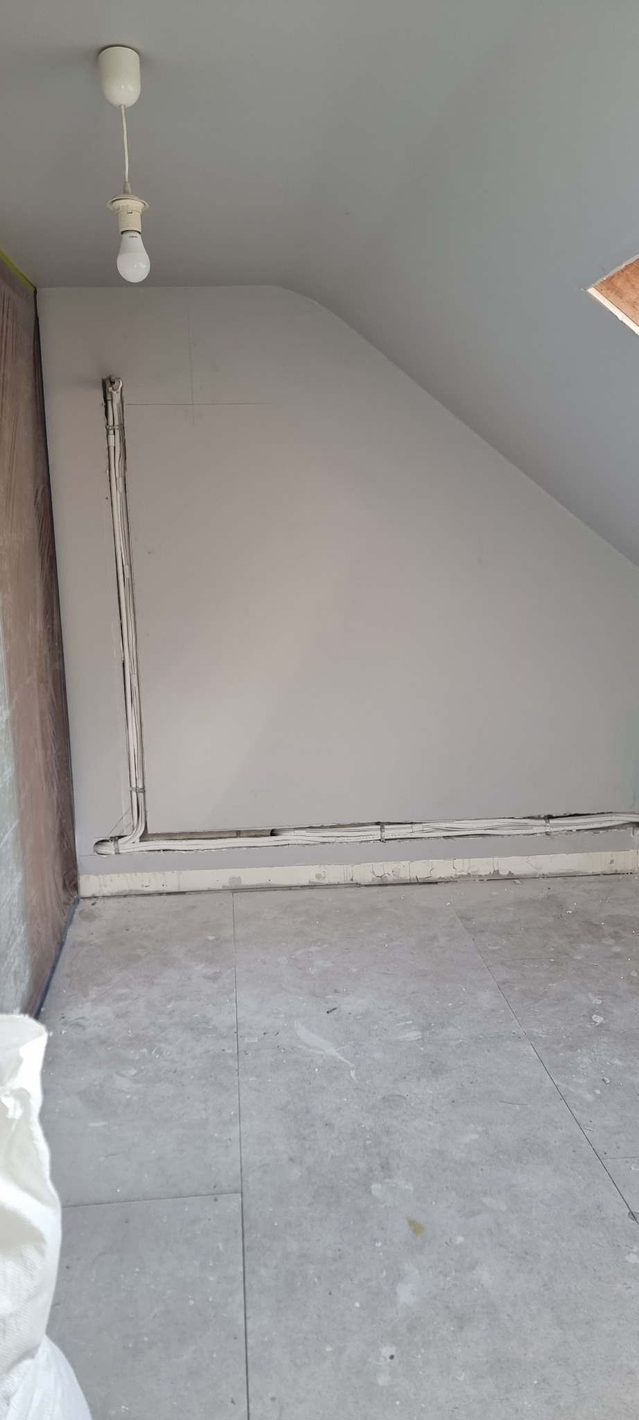 Instalacja elektryczna w szarej wnęce poddasza. Kable ułożone wzdłuż ściany i podłogi, mocowane uchwytami. Widoczna żarówka na kablu.