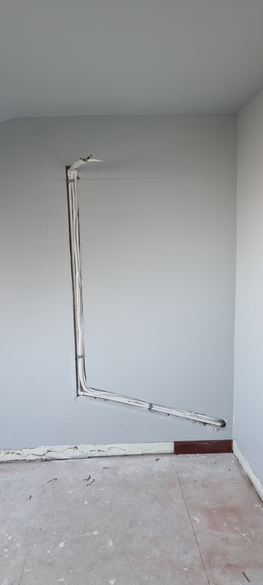 Białe przewody elektryczne ułożone w bruździe w szarej ścianie podczas remontu. Widoczne mocowania kabli i fragment podłogi z listwą przypodłogową.