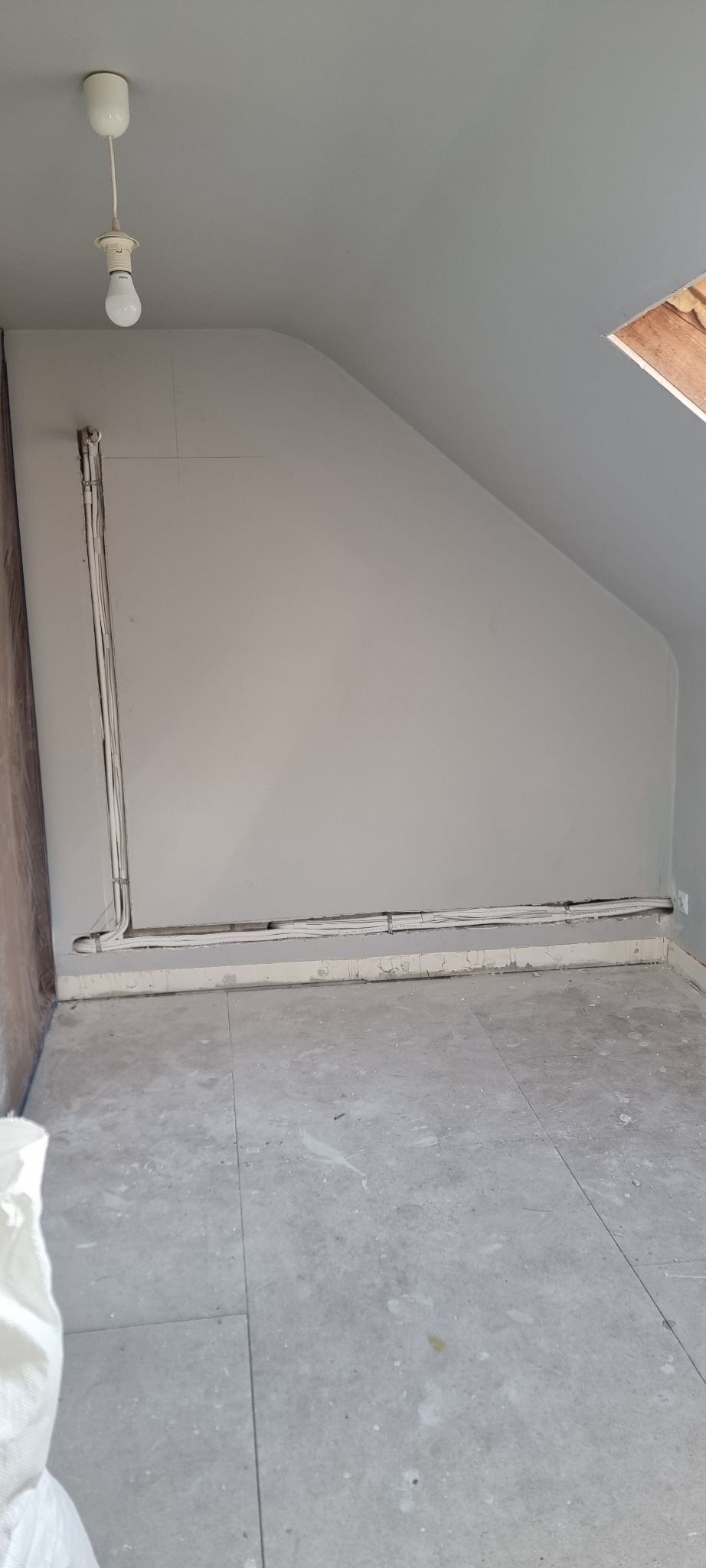 Poddasze w trakcie remontu z widocznymi przewodami elektrycznymi ułożonymi wzdłuż ściany i pod sufitem, surowy beton na podłodze, widok na okno dachowe.