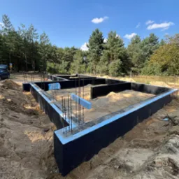 Fundamenty domu jednorodzinnego w trakcie budowy, z widoczną izolacją przeciwwilgociową i zbrojeniami, otoczone piaskiem, las w tle, słoneczny dzień.