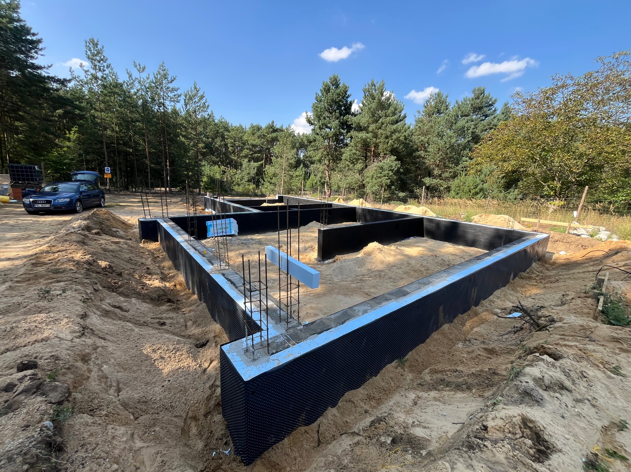 Fundamenty domu jednorodzinnego w trakcie budowy, z widoczną izolacją przeciwwilgociową i zbrojeniami, otoczone piaskiem, las w tle, słoneczny dzień.