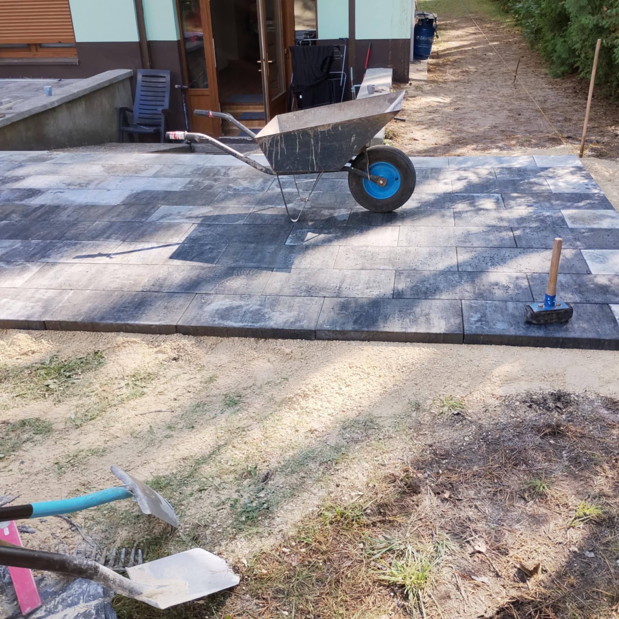 Ułożona kostka brukowa z wózkiem i narzędziami na piasku. Widoczne szare, prostokątne płyty i przygotowany teren. Praca w toku, widok z góry.