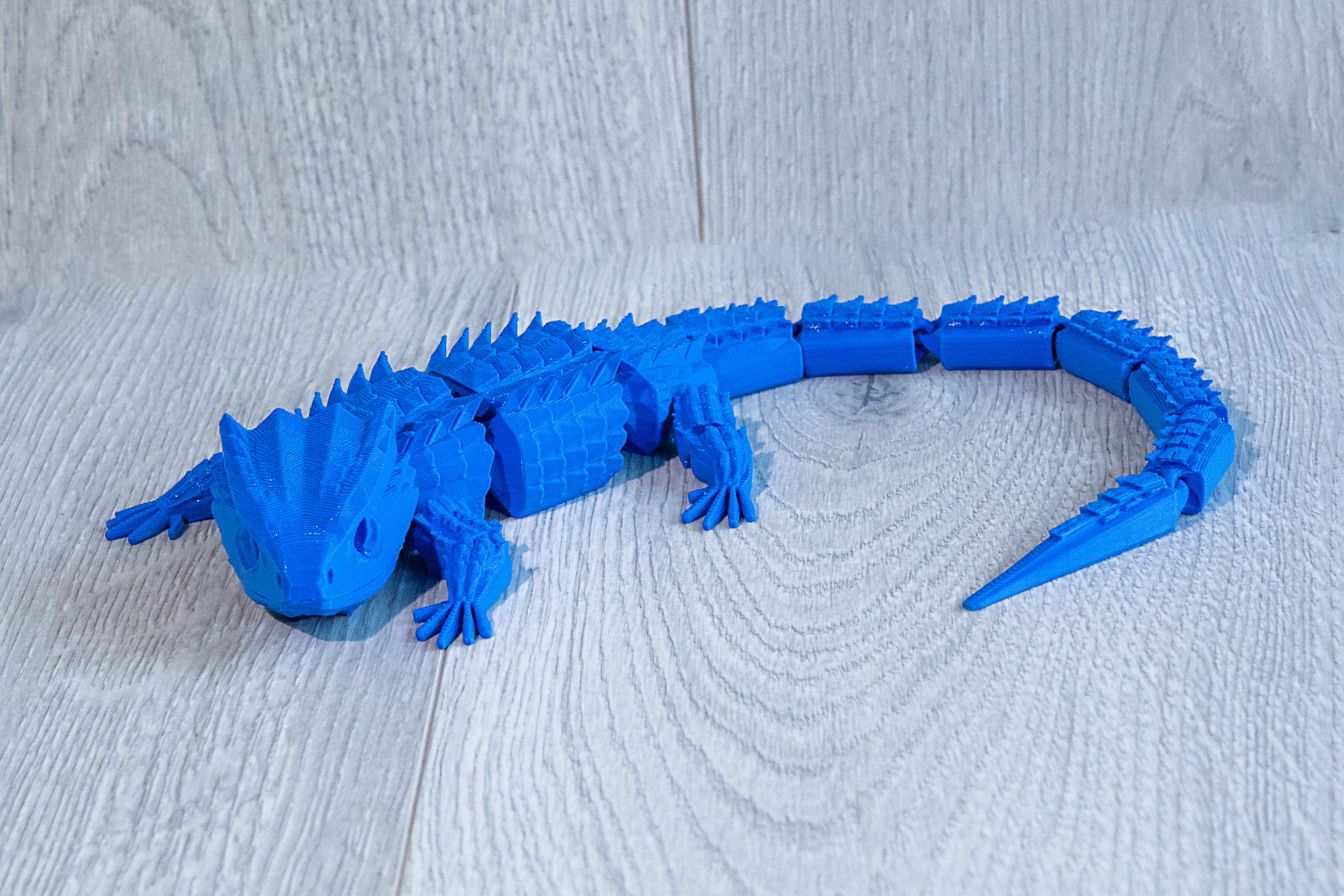 Niebieska, ruchoma figurka jaszczurki wydrukowana w 3D, leżąca na drewnianym tle. Widoczne warstwy druku, imitujące łuski. Precyzyjne odwzorowanie detali.