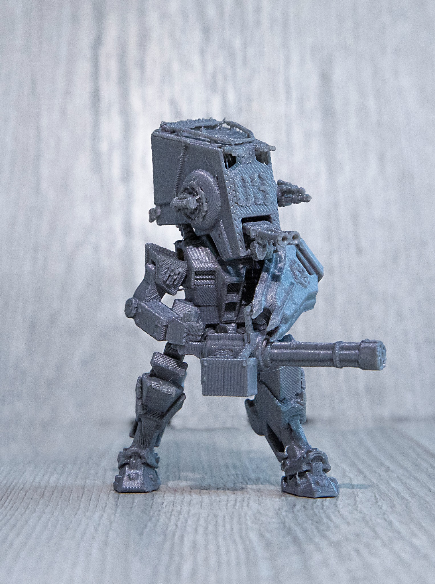 Szary model mecha z drukarki 3D, z karabinem maszynowym, na szarym tle. Widoczne warstwy druku, szczegółowy projekt.