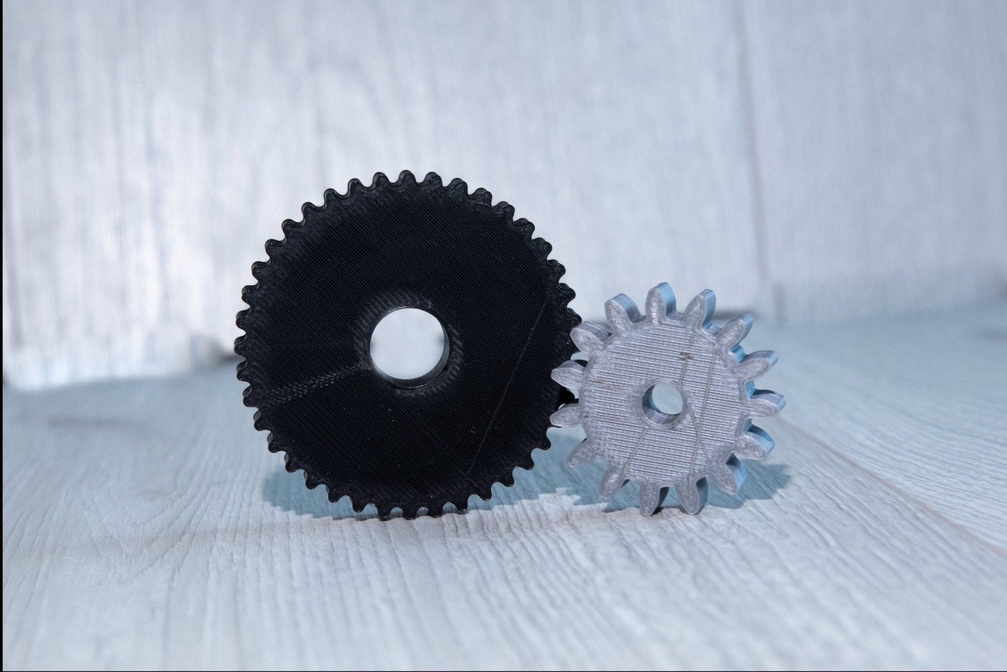 Dwie plastikowe zębatki: czarna, większa i srebrna, mniejsza, wydrukowane w 3D. Widoczne warstwy druku, ustawione na jasnym, drewnianym tle.