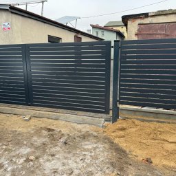 OGROTKOWO MARCIN GROTEK - Nowoczesna, pozioma brama wjazdowa z szarych paneli na tle budynku z panelami słonecznymi. Brama częściowo otwarta, widoczny piasek i elementy betonowe w podłożu.