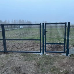 OGROTKOWO MARCIN GROTEK - Nowoczesna, grafitowa brama z siatki panelowej, zamontowana na betonowej podmurówce, na tle zamglonego krajobrazu z drzewami. Widoczne elementy montażowe.