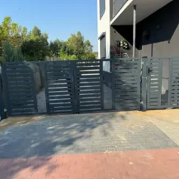 Nowoczesna, grafitowa brama wjazdowa z poziomymi szczeblami, uzupełniona kratką. Brama zamontowana na posesji z kostką brukową, z domem w tle.