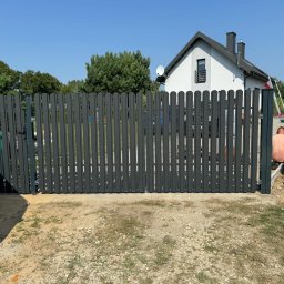 OGROTKOWO MARCIN GROTEK - Grafitowa brama wjazdowa z pionowych sztachet, częściowo zamontowana. W tle dom z białą elewacją i ciemnym dachem, fragment sylwetki pracownika.