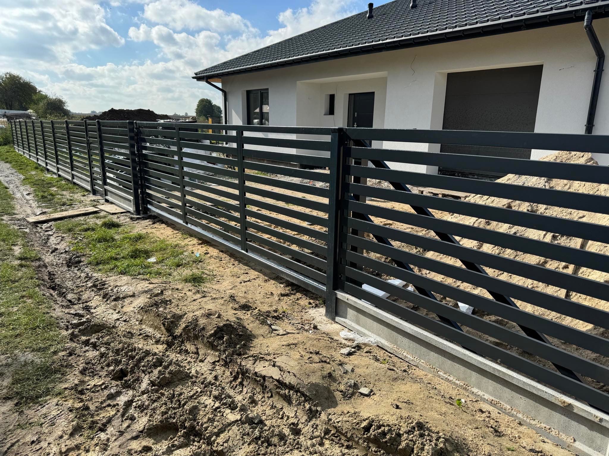 Nowoczesne, poziome ogrodzenie metalowe w kolorze grafitowym, z betonową podmurówką, na tle białego domu z grafitowym dachem w słoneczny dzień.