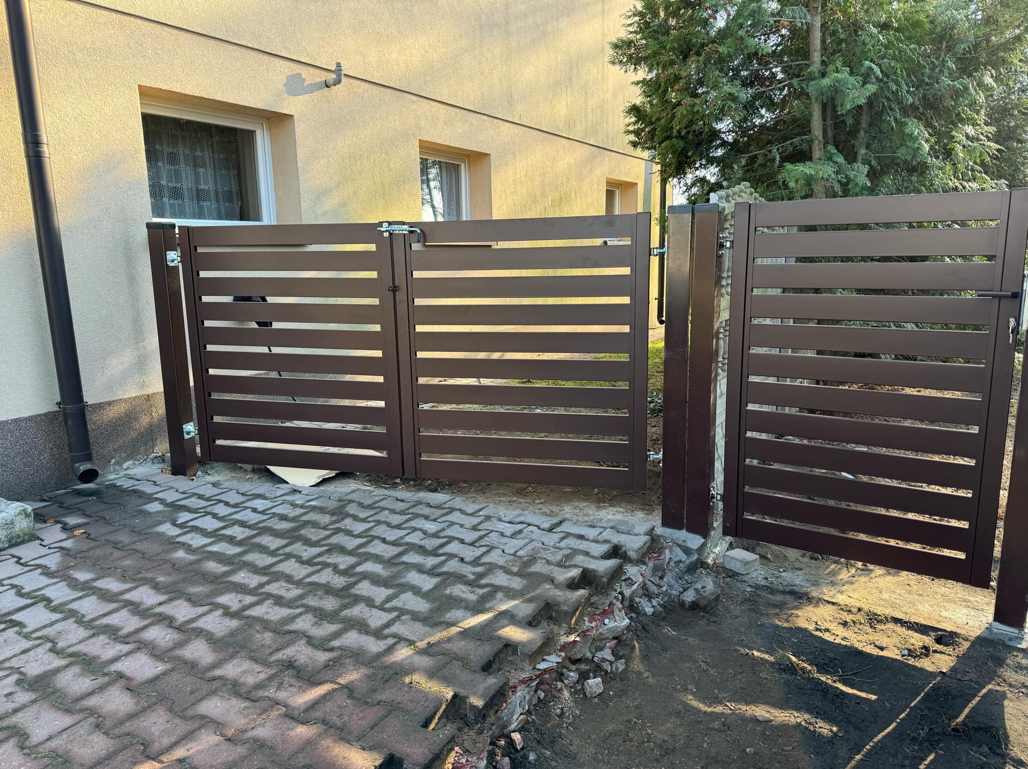 Brązowa, nowoczesna brama wjazdowa z poziomymi panelami, zamontowana na posesji z kostką brukową. Widoczne ślady prac ziemnych.