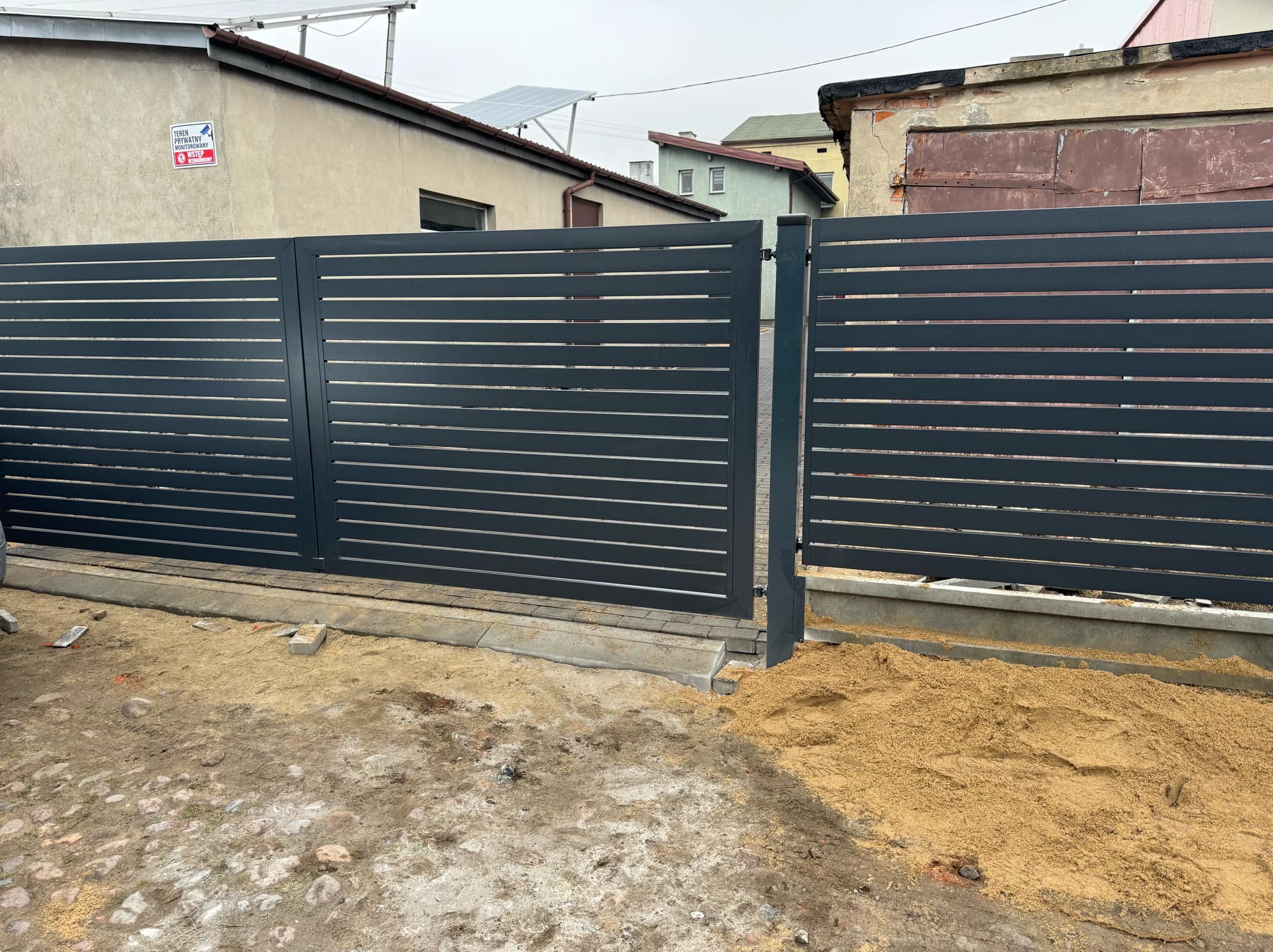 Nowoczesna, pozioma brama wjazdowa z szarych paneli na tle budynku z panelami słonecznymi. Brama częściowo otwarta, widoczny piasek i elementy betonowe w podłożu.