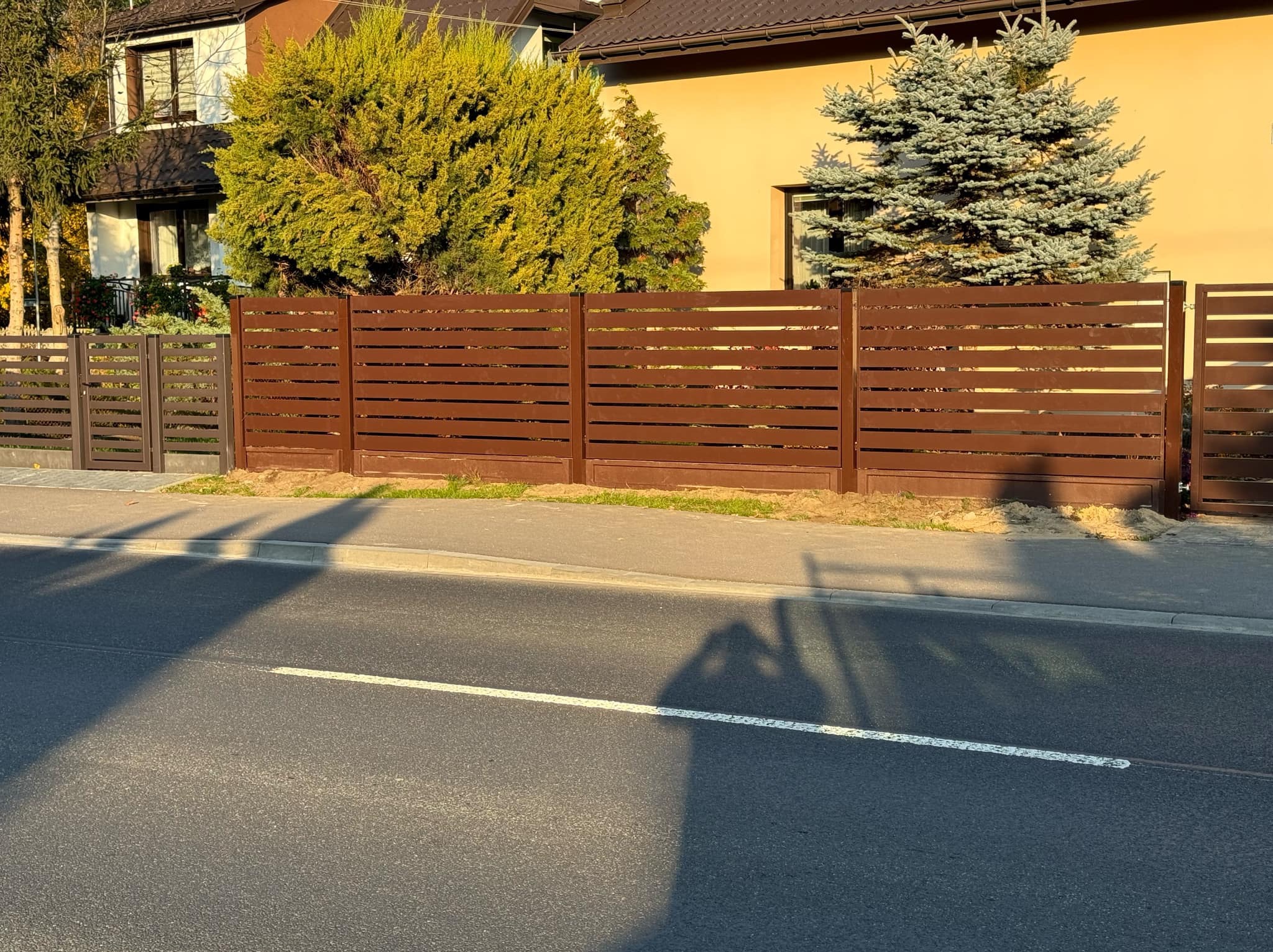 Nowoczesne, brązowe ogrodzenie panelowe z poziomymi szczeblami, oddzielające posesję od ulicy. W tle dom jednorodzinny i drzewa.