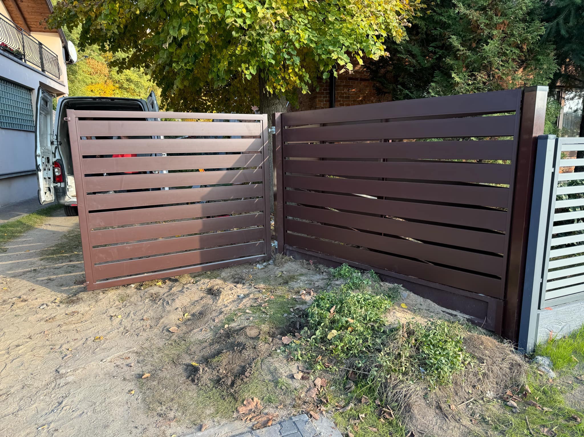 Otwarta, brązowa brama wjazdowa z poziomych profili, widoczna na tle zieleni i domu. Brama częściowo zamontowana, obok zaparkowany biały bus.