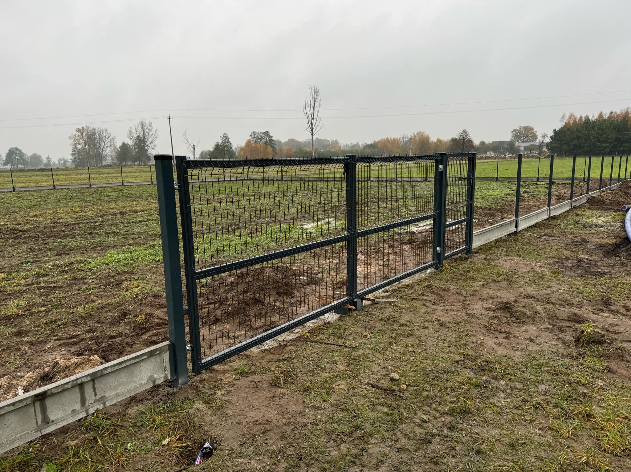 Szara, metalowa brama z siatki, część ogrodzenia, na betonowej podmurówce. Widok na otwarty teren z drzewami w tle. Pochmurny dzień.